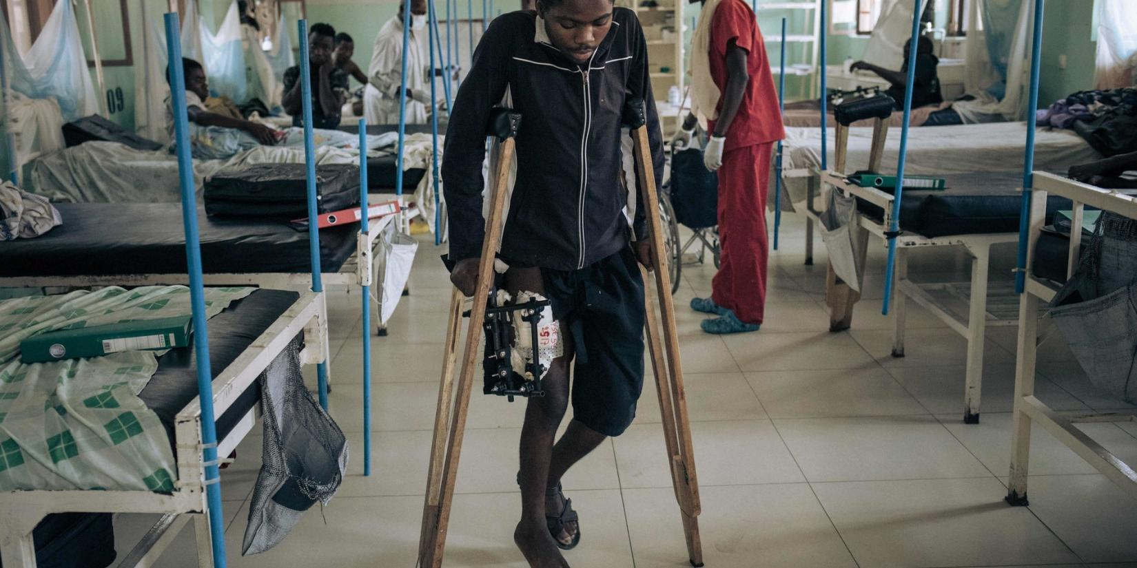 A young African man with a gunshot wound to the leg walks with difficulty on crutches in a hospital.