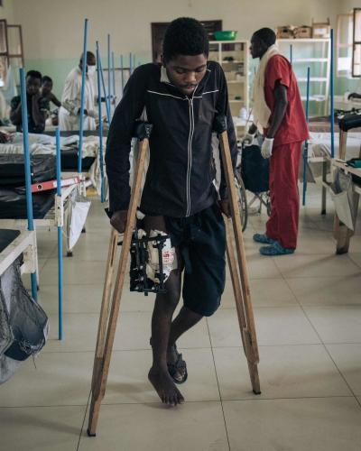 A young African man with a gunshot wound to the leg walks with difficulty on crutches in a hospital.